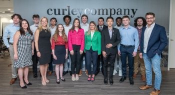 A group of professionally dressed students and mentors stand together indoors in front of a wall with the Curley Company logo.