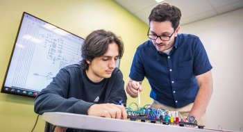 A student and instructor work together on a circuit board project, with a wiring diagram displayed on a screen behind them. The student focuses on the components, while the instructor gestures with a stylus. They are in a brightly lit classroom with light green walls.