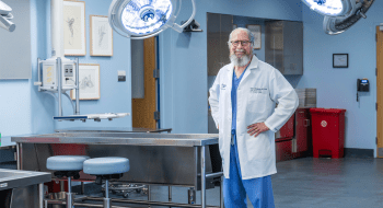 Robert Montgomery, wearing blue scrubs and a white lab coat, stands in a hospital operating room. Three are large florescent lights above him.