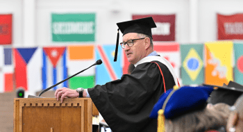Michael Ranger, wearing graduation regalia, stands at a podium and address the crowd. Several countries flags are in the background.