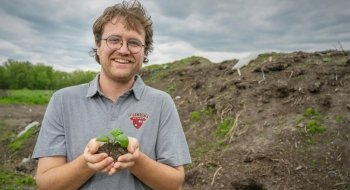 Sam Voter, wearing a grey Saint Lawrence University polo, holds a handful of dirt with a green sprout in the center, a large compositing pile is in the background.