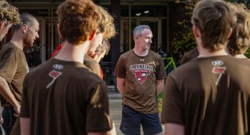 Paul Graham wearing a &quot;Saint Lawrence Cross Country&quot; shirt stands in the center of a group of team members during an outdoor meeting.