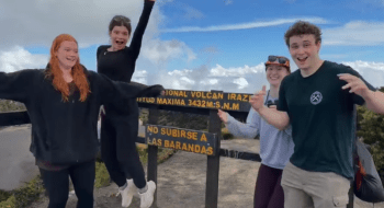 Four smiling students pose and celebrate in front of a sign at Irazú Volcano National Park in Costa Rica, with one student mid-jump. The sign shows an elevation of 3,432 meters above sea level. The sky is partly cloudy with scenic views in the background.