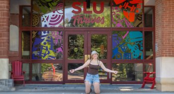 Student kneels and smiles in front of the Sullivan Student Center at Saint Lawrence University, with colorful window art and the words "SLU Thrives" above the entrance.