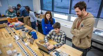 Students collaborate at a workbench in a Saint Lawrence University lab, using a laptop and electronic components. One student types while others stand nearby, observing and discussing the project. Tools, wires, and equipment are spread across the table, showing hands-on learning in progress.