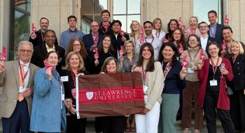 St. Lawrence University alumni and staff pose on the steps of a historic building, holding a university banner and red foam fingers.