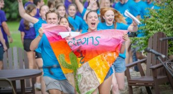 Students cheer while carrying a colorful handmade banner during an outdoor campus event.