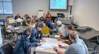 Students work together on a large group activity at classroom tables while a facilitator gives instructions at the front of the room.