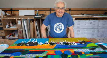 Woody Jackson, wearing a blue T-shirt stands behind a brightly colored painting featuring abstract shapes, bold stripes, and black-and-white cows in an art studio.