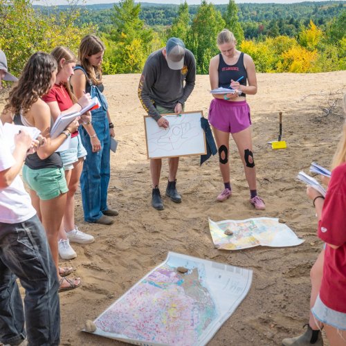 A group of Saint Lawrence University students stands in a circle outdoors, observing a peer drawing on a whiteboard with maps spread on the ground.