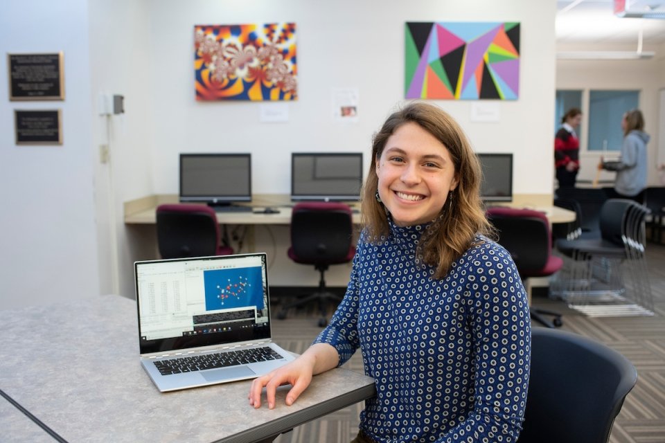 A photo of alum Nicole Panek who is smiling and sitting by her laptop that contains many pieces of data.