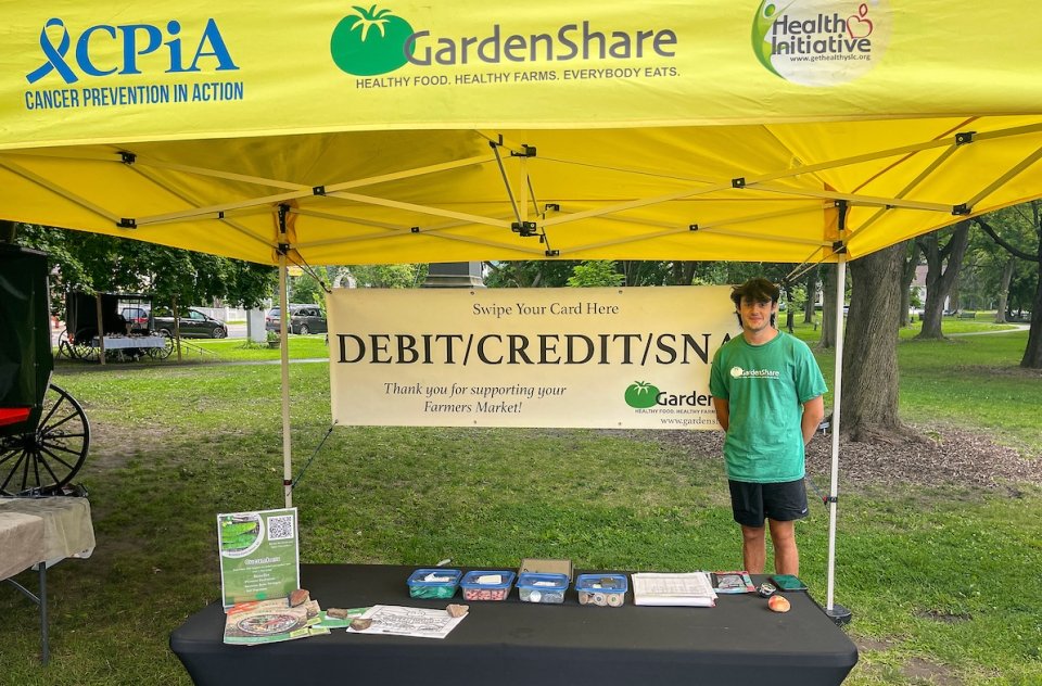 Ryan Krugman stands next to a sign that reads "Debit, Credit, SNAP" under a yellow tent, representing GardenShare.