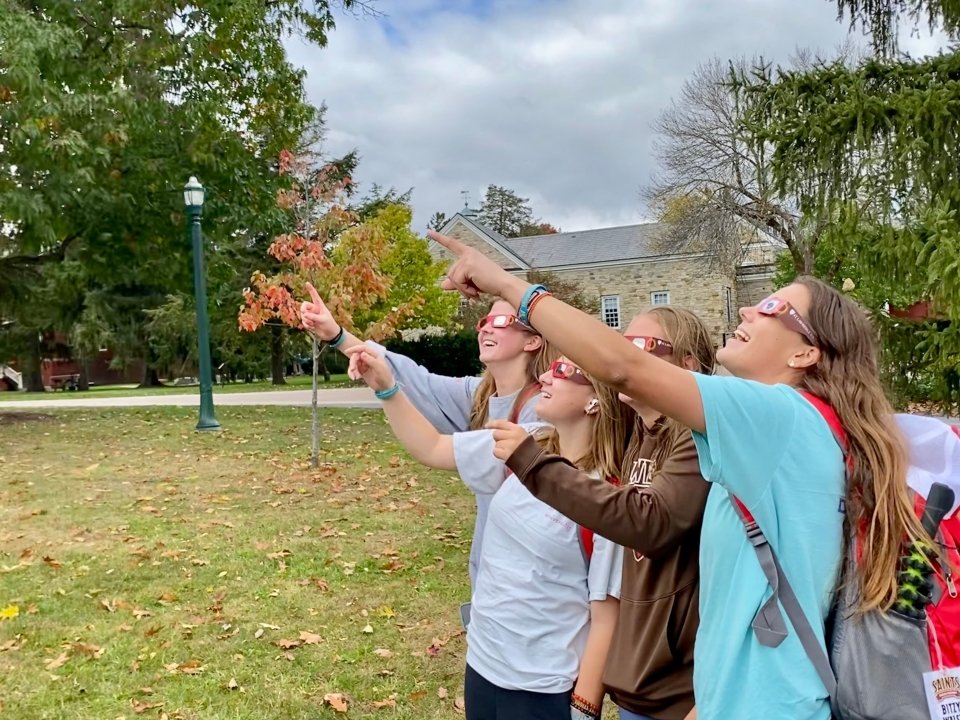 Four young women wearing eclipse glasses point at the partial solar eclipse over campus.