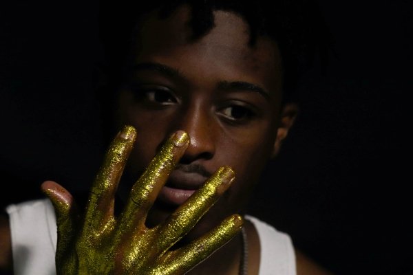 Kaleb Davis models for a photo series titled "Forbidden Crowns." He wears a white tank top and holds a hand covered in gold glitter up to his face.