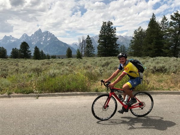 Phil Duggan riding a bike past a grassy, open field with the Teton Mountains towering in the background. 