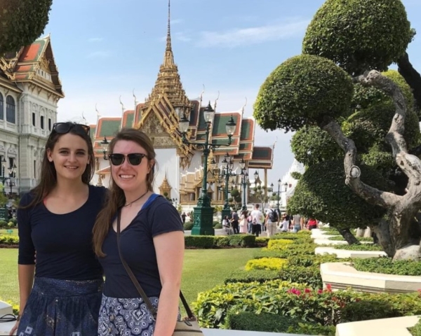 Two young women stand in front of a verdant lawn and an elegant, elaborate temple in Thailand.