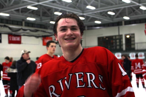 Tommy Benjes, wearing a red "Rivers" hockey jersey, smiles wide while skating off the ice.