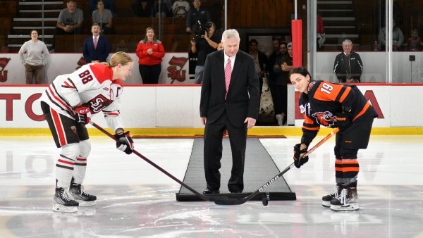 Joe Marsh performs a ceremonial puck drop as two women's ice hockey players prepare for the drop.