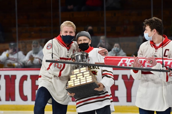 Sophomore Greg LaPointe, wearing a Saints hockey jersey and a knitted cap, holds a large hockey championship trophy while posing for a photo with his teammate on the ice in Appleton Arena.
