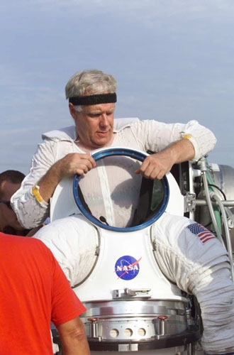 Dean Eppler, wearing a black headband, hoists up a large, white NASA space suit.