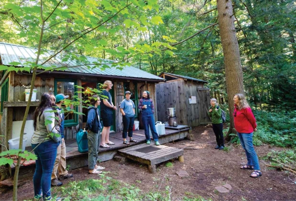 President Morris, wearing a scarlet jacket, stands with a group of students in the woods and listens to their experience living off-the-grid.
