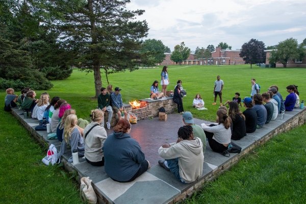 A group of first-year students sit on a stone patio around a fire, listening to their Orientation Leader. In the background, is a large grassy lawn and a college dorm building.