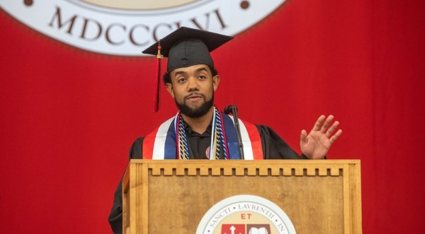 Brian Uceta, wearing academic regalia, stands at a podium and addresses the crowd during Commencement 2023. A scarlet banner with the Saint Lawrence crest is in the background.