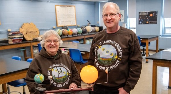 Aileen O'Donoghue and Jeffrey Miller wear brown Saint Lawrence University Gateway to the Path of Totality sweatshirts and stand in a physics lab.