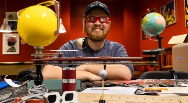 Tyler Karasinksi, wearing red solar eclipse glasses, crosses his arms on top of a desk that is covered in eclipse memorabilia and model of the solar system.