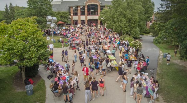 A bustling outdoor event on the Saint Lawrence University campus with rows of tables, students, and community members engaging. The scene is surrounded by trees and buildings, with colorful booths and activities along the pathways.