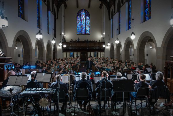 Orchestra performs in a chapel while a conductor leads, with a large audience seated in pews beneath stained glass windows.