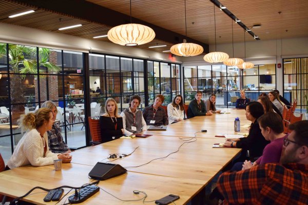A group of students and adults seated around a large wooden conference table during a meeting in a modern, glass-walled room with pendant lighting..