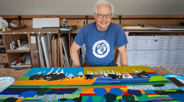 Woody Jackson, wearing a blue T-shirt stands behind a brightly colored painting featuring abstract shapes, bold stripes, and black-and-white cows in an art studio.