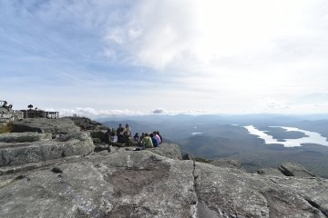 A class gathers on a rocky ledge at the summit of a mountain overlooking the Adirondack mountains and a large lake.