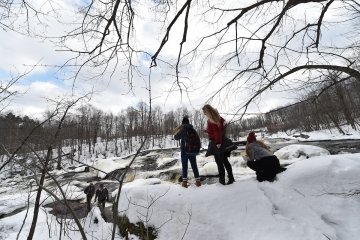 A wintery outdoor scene. Three students stand on a snow-covered trail overlooking a waterfall.