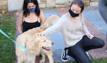 Two students, both wearing masks, kneel on the grass while petting a golden retriever.
