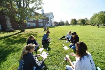 A group of students sit cross-legged on the grass while working on a painting project.