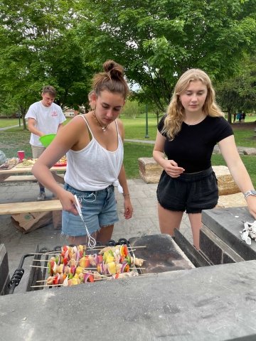 Two female students grilling kabobs with trees and a picnic table in the background.