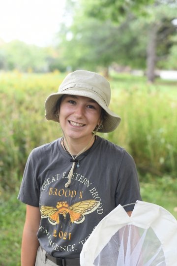 Karina Bellavia, wearing a bucket hat and holding an insect net, stands in front of a wildflower field.