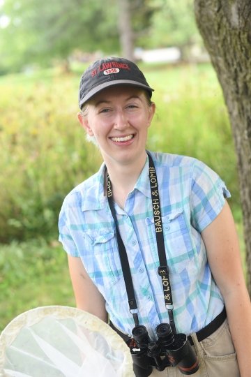 Kayla Edmunds, wearing a gray Saint Lawrence hat and holding an insect net, stands in front of a wildflower field. There are binoculars around her neck.