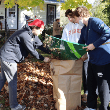 Three Saint Lawrence University students hold out a large brown paper bag and collect leaves as part of a community service project. 