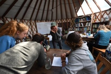 A professor guides a lively, hands-on class session for students who gather around a microscope at the table in front of them. They are inside an Adirondack yurt classroom.