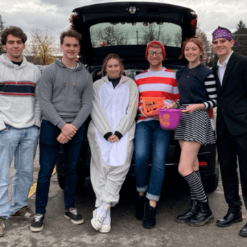 Saint Lawrence students, Hallie Heggeness, Tyler Kearns, Alexandra Hill, and Trent Meyer, and other college students stand at the trunk of a car ready to hand out Halloween candy.