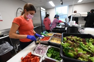 A student, wearing a mask and disposable gloves, prepares a healthy meal with fresh veggies.