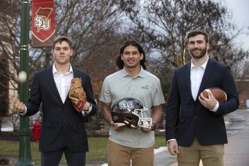 Andrew Matthews, Tyler Grochet, and Ben Barba, wearing business attire and hold athletic equipment, stand next to one another on a campus walkway on an overcast day.