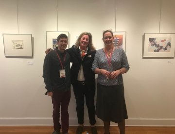 Brendan Reilly, Abby Daly, and Melissa Schulenberg at an art gallery exhibit. Framed artwork hangs on the wall behind them. 