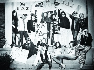 A black and white photo of old Kappa Delta Sigma members on the front steps of their house all holding KDS signs.