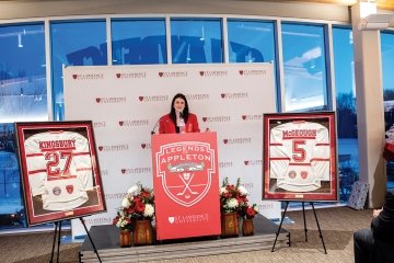 A photo of alumni Gina Kingsbury speaking behind a podium with two framed jerseys behind her.