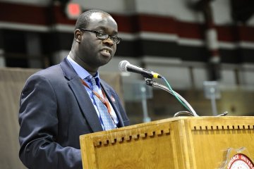 A photo of Okoth standing behind a podium. 