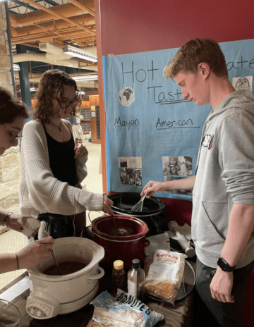Three student gather around crock pots, stirring liquid chocolate for a hot cocoa tasting. 